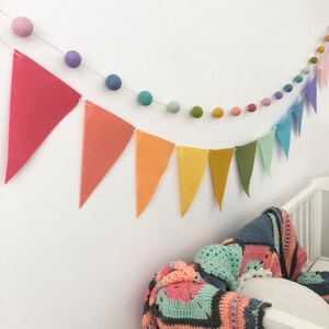 May include: A colorful nursery decoration featuring two garlands and a crocheted blanket. One garland has felt pennants in a rainbow of colors, and the other has felt balls. The blanket is a patchwork of various colors and patterns.