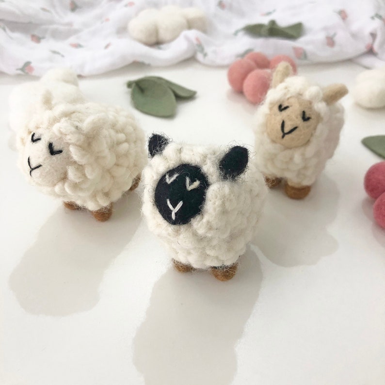 May include: Three white felt sheep toys with embroidered faces. One sheep has black felt eyes and a black nose. The other two sheep have black embroidered eyes and noses.