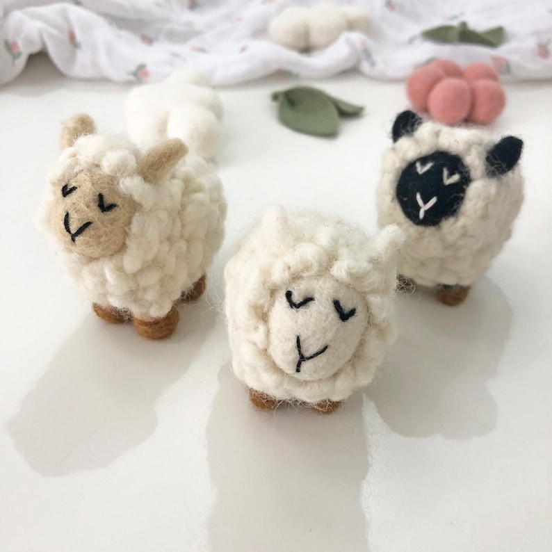 May include: Three white felt sheep with black embroidered eyes and mouths. The sheep are standing on a white surface.
