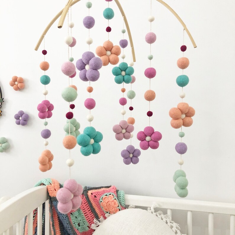 May include: A hanging mobile featuring felted wool balls and flower-shaped felt pieces in shades of lavender, peach, mint green, and white. The elements are suspended from a curved wooden frame by thin white strings, creating a colorful display.