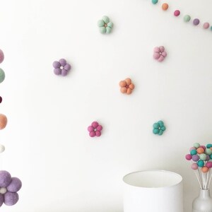 May include: A collection of felted wool flower decorations in various pastel colors including mint, lavender, peach, and pink, arranged individually on a wall alongside a felt ball garland and a bouquet of felted spheres on white stems.