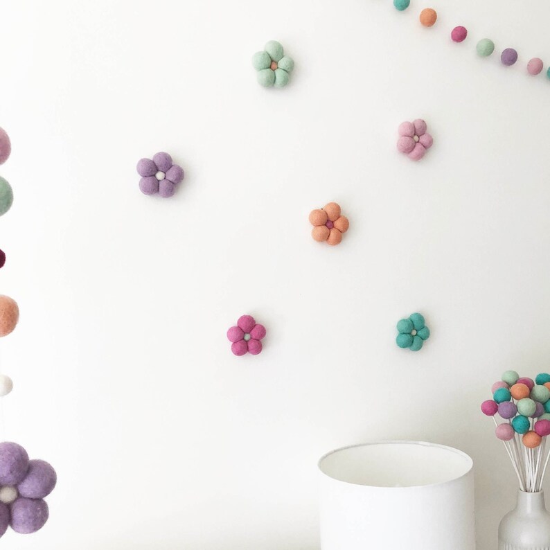 May include: A collection of felted wool flower decorations in various pastel colors including mint, lavender, peach, and pink, arranged individually on a wall alongside a felt ball garland and a bouquet of felted spheres on white stems.