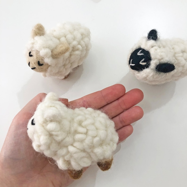 May include: Three white wool felt sheep, one with black eyes and nose, one with brown eyes and nose, and one with black eyes and nose. The sheep are all lying down.