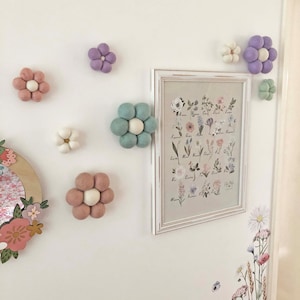 May include: A collection of felt flower decorations in various pastel colors, including pink, purple, and green, are affixed to a white wall. A framed botanical print and floral decals add to the decorative theme. The flowers are arranged in a playful, whimsical style.