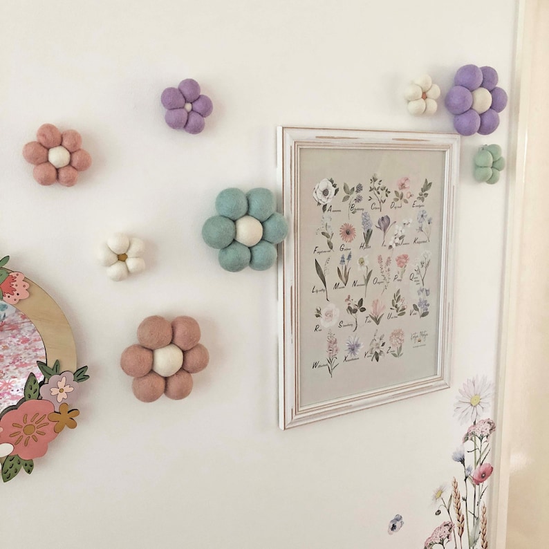 May include: A collection of felt flower decorations in various pastel colors, including pink, purple, and green, are affixed to a white wall. A framed botanical print and floral decals add to the decorative theme. The flowers are arranged in a playful, whimsical style.