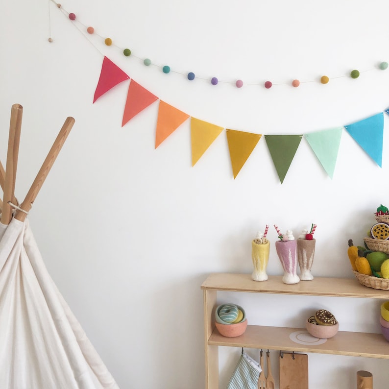 May include: A playroom scene with a white teepee and a wooden shelf. A rainbow-colored bunting and felt ball garland decorate the wall. The shelf displays toy milkshakes, donuts, and a basket of toy fruits and vegetables.
