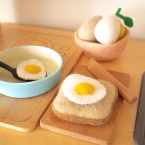 May include: A felt play food set featuring a light blue pan with a fried egg, a slice of toast topped with a fried egg, a wooden knife, and a small bowl containing two felt eggs, one white and one beige, with a small green felt leaf.