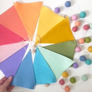 May include: A colorful felt banner with triangular flags in shades of orange, yellow, green, blue, and purple. A string of felt balls in complementary colors is also present. The banner is perfect for decorating a nursery or children's room.