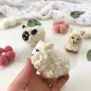May include: Three white felt sheep toys with black eyes and brown legs. The sheep are on a white surface with pink felt flowers.
