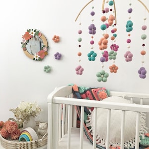 May include: A nursery mobile featuring felt balls and flower shapes in shades of teal, peach, lavender, and white hanging from a wooden frame. A circular mirror with a floral wooden frame and three small felt flowers are mounted on the wall.