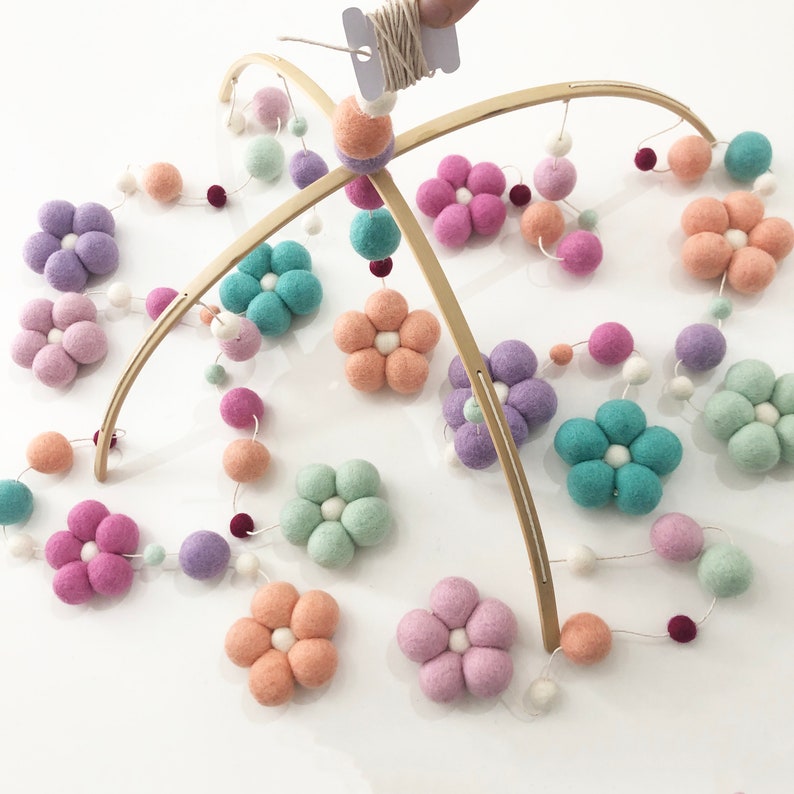 May include: A hanging mobile featuring curved wooden arches and strings of felted wool elements. The design includes multi-colored felt flowers in shades of lavender, peach, teal, and mint, interspersed with small white, purple, and dark red felt balls.