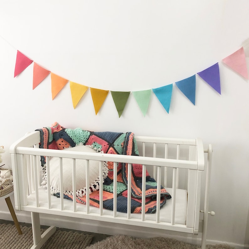 May include: A white crib with a colorful crochet blanket and a white pillow. A rainbow-colored pennant banner hangs above the crib. The blanket features squares of pink, blue, and green.