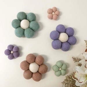 May include: A collection of felt flower decorations in various colors, including green, purple, pink, and brown. Each flower is composed of multiple felt balls arranged in a floral pattern, with a white center. The flowers are mounted on a white surface.