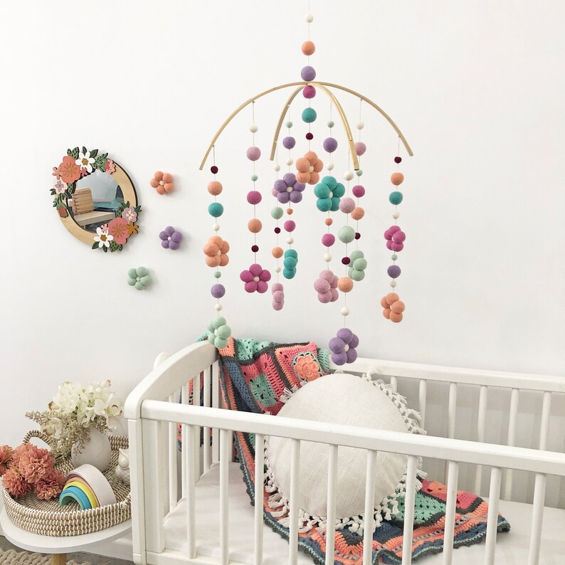 May include: A hanging mobile featuring felt balls and felt flowers in shades of peach, lavender, teal, and mint green suspended from a curved wooden frame. The mobile includes multiple strands of varying lengths with small white beads between the felt elements.