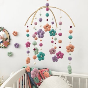 May include: A hanging mobile featuring felted wool balls and flower-shaped felt pieces in shades of lavender, peach, mint green, and teal. The elements are suspended from two intersecting curved wooden arches by thin white strings.