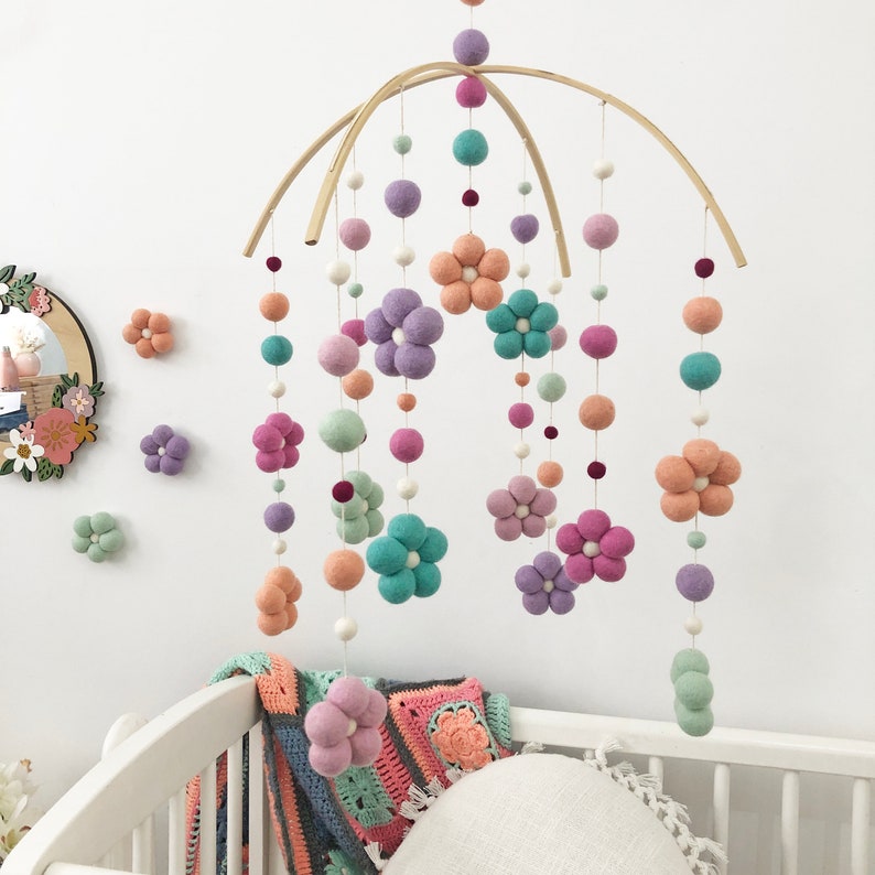 May include: A hanging mobile featuring felted wool balls and flower-shaped felt pieces in shades of lavender, peach, mint green, and teal. The elements are suspended from two intersecting curved wooden arches by thin white strings.