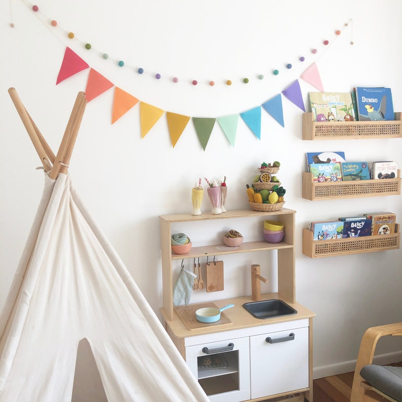 May include: A children's playroom with a white teepee, a wooden play kitchen, and rattan bookshelves. Colorful pennant banners and felt ball garlands decorate the white walls. The kitchen has a sink, stove, and storage. Books and toys are arranged throughout the room.