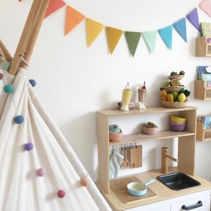 May include: A child's play kitchen set with a wooden frame, featuring a sink, stove, and storage shelves. The set is decorated with toy food, bowls, and utensils. A colorful garland hangs above, and a teepee is on the left.