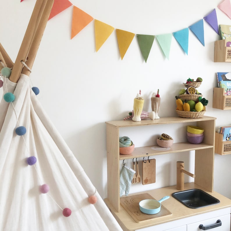 May include: A child's play kitchen set with a wooden frame, featuring a sink, stove, and storage shelves. The set is decorated with toy food, bowls, and utensils. A colorful garland hangs above, and a teepee is on the left.