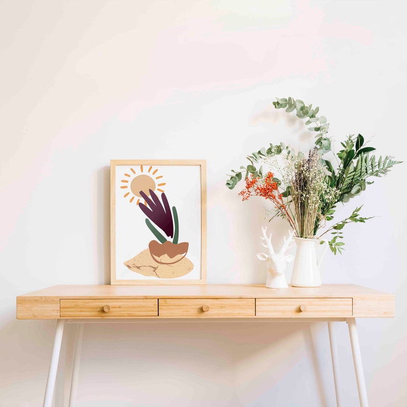 May include: A framed print of an abstract art piece with a sun, a plant, and a brown pot on a light beige background. The print is on a wooden desk with a white vase and a white deer figurine.