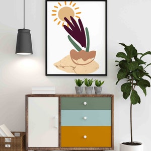 May include: A framed abstract art print with a sun, a plant, and a brown and beige background. The print is hanging on a white wall above a wooden dresser with three drawers.