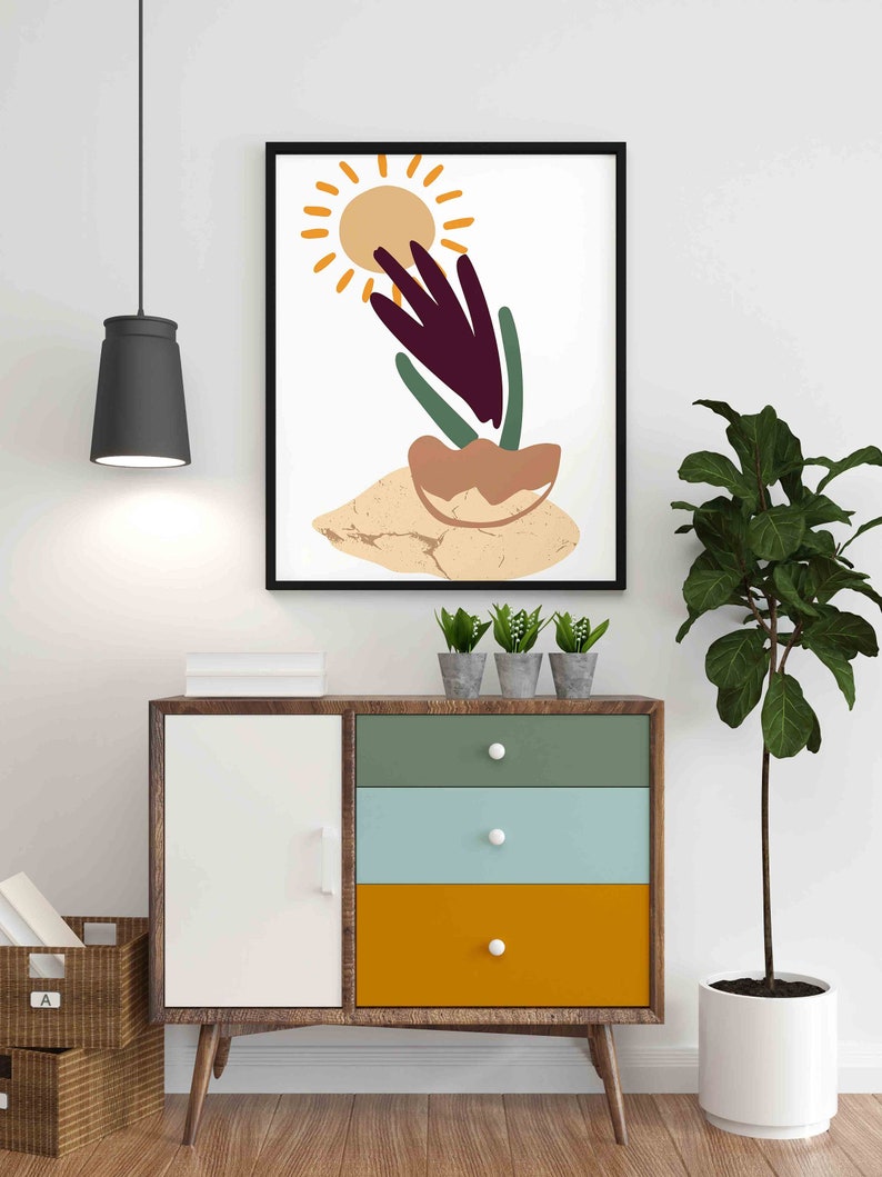 May include: A framed abstract art print with a sun, a plant, and a brown and beige background. The print is hanging on a white wall above a wooden dresser with three drawers.