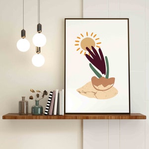 May include: A minimalist art print with a stylized sun, a purple plant, and a brown pot on a beige background. The print is framed in a black frame and is hanging on a white wall above a wooden shelf.
