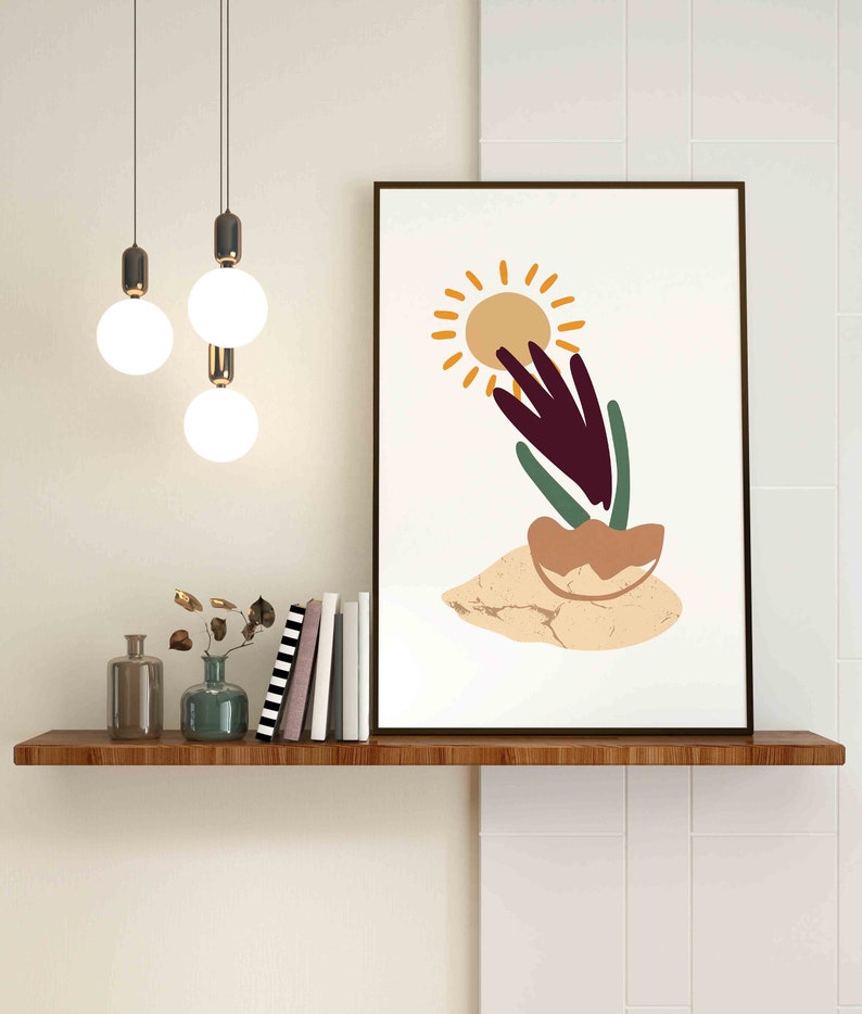May include: A minimalist art print with a stylized sun, a purple plant, and a brown pot on a beige background. The print is framed in a black frame and is hanging on a white wall above a wooden shelf.