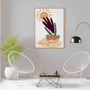 May include: A framed abstract art print with a sun, a plant, and a brown rock on a beige background. The print is hanging on a light gray wall above a coffee table with a chessboard and two white chairs.
