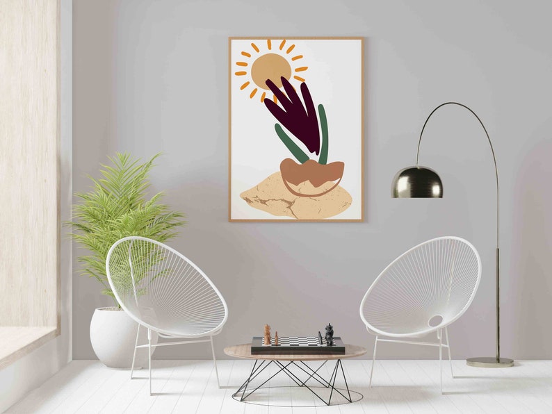 May include: A framed abstract art print with a sun, a plant, and a brown rock on a beige background. The print is hanging on a light gray wall above a coffee table with a chessboard and two white chairs.