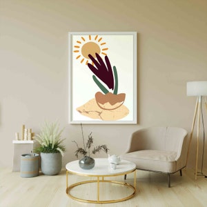 May include: Abstract art print featuring a stylized sun, a plant with green leaves and a purple stem, and a brown pot on a beige background. The print is framed in white and hangs on a beige wall.