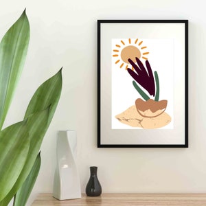 May include: A black framed print with a white background featuring a stylized illustration of a sun, a plant with purple and green leaves, and a brown rock. The illustration is in a minimalist style.