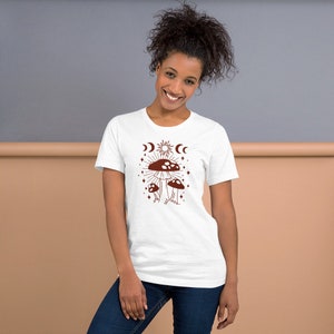 Könnte beinhalten: Weißes T-Shirt mit einem braunen Pilz-Motiv. Der Pilz ist von einer Sonne, einem Mond und Sternen umgeben. Der Text "Shroom Queens" ist auf das T-Shirt gedruckt.