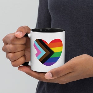 May include: A white ceramic mug with a black rim and handle. The mug features a heart-shaped design incorporating the colors of the Progress Pride flag.
