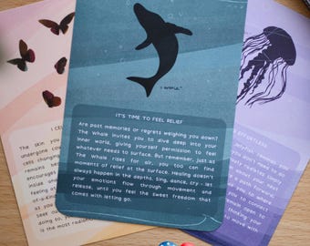 Puede incluir: Tarjetas de oráculo con ilustraciones y texto. Una tarjeta presenta una silueta de ballena, otra una medusa y una tercera con mariposas. Las tarjetas contienen mensajes inspiradores sobre la autorreflexión y la liberación emocional.