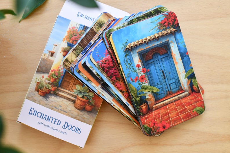 May include: An oracle deck titled "Enchanted Doors" with a stack of cards featuring colourful illustrations of doors and floral arrangements. The box and cards are on a wooden surface. The cards have rounded corners.