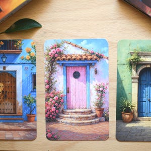 May include: Three rectangular art prints depicting watercolour illustrations of doors. One shows a blue building with a wooden door, another a pink door with flowers, and the last a green building with a blue door.