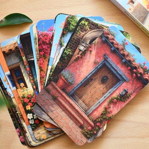 May include: A deck of oracle cards titled 'Enchanted Doors' fanned out on a wooden surface. Each card displays a colourful illustration of a door or building facade with flowers and architectural details. A box with the same title is visible.