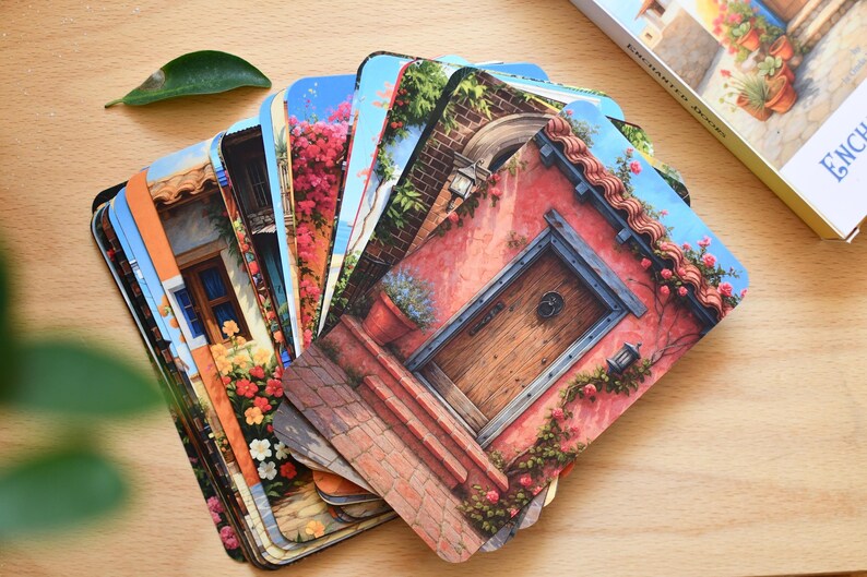 May include: A deck of oracle cards titled 'Enchanted Doors' fanned out on a wooden surface. Each card displays a colourful illustration of a door or building facade with flowers and architectural details. A box with the same title is visible.