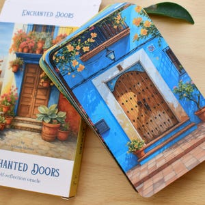 May include: A deck of "Enchanted Doors" self-reflection oracle cards. The cards and box feature a watercolour illustration of a blue building with a wooden door, flowers, and potted plants. The box has the text "Enchanted Doors".