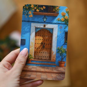 May include: A rectangular art print showing a blue building facade with a wooden door. The building has a balcony with yellow flowers and potted plants. The door is brown with decorative studs, and the scene is bathed in sunlight.
