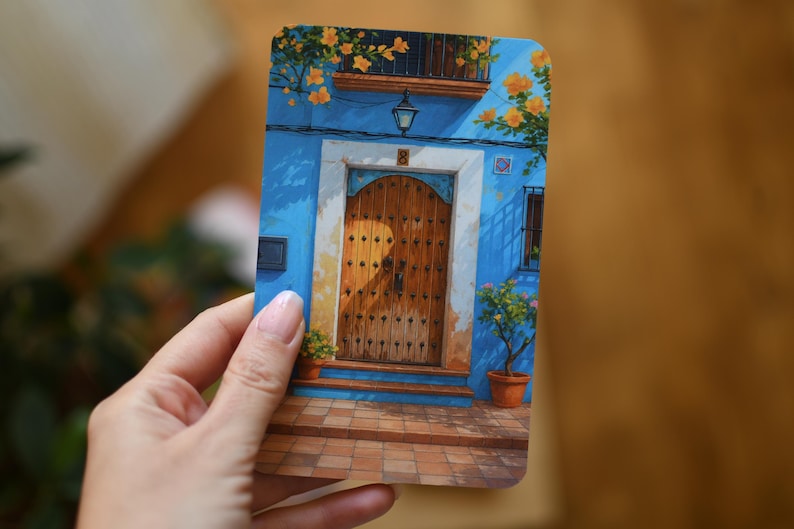 May include: A rectangular art print showing a blue building facade with a wooden door. The building has a balcony with yellow flowers and potted plants. The door is brown with decorative studs, and the scene is bathed in sunlight.