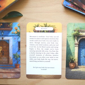 May include: Three oracle cards with watercolour illustrations of doorways and buildings. The cards feature blue, green, and yellow hues, with text on one card about movement. The cards are on a wooden surface.