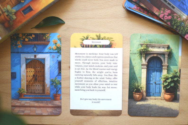 May include: Three oracle cards with watercolour illustrations of doorways and buildings. The cards feature blue, green, and yellow hues, with text on one card about movement. The cards are on a wooden surface.