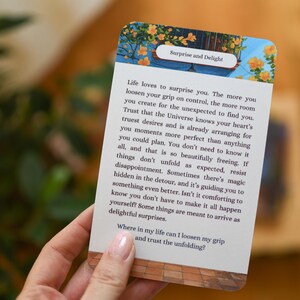 May include: A card with the text "Surprise and Delight" and a message about life's surprises. The card has a floral design at the top and is held by a person. The background is blurred.