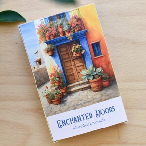 May include: A rectangular box with a watercolour painting of a colourful building with a blue door, potted plants, and the text "ENCHANTED DOORS self-reflection oracle". The box is on a light brown wooden surface.