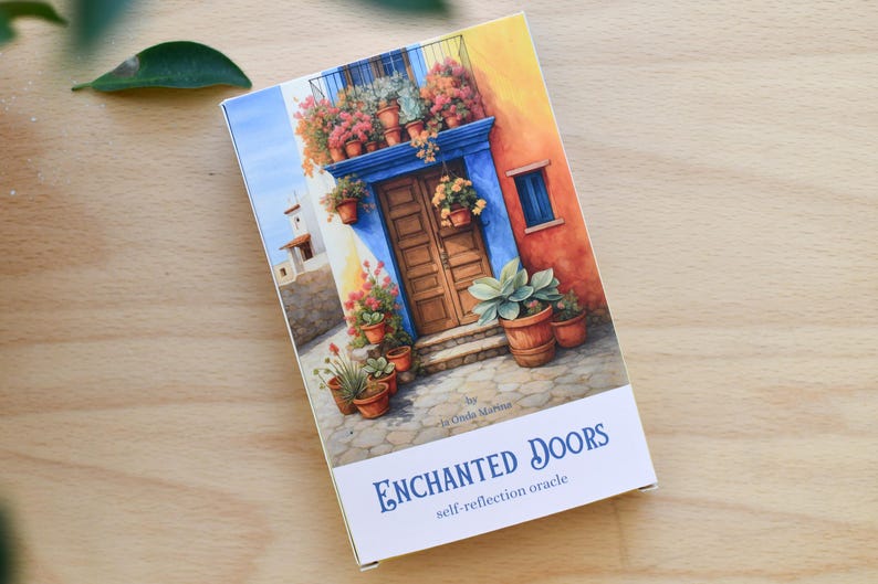 May include: A rectangular box with a watercolour painting of a colourful building with a blue door, potted plants, and the text "ENCHANTED DOORS self-reflection oracle". The box is on a light brown wooden surface.