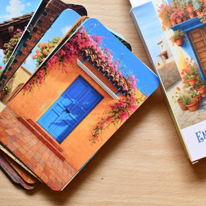 May include: A deck of Enchanted Doors self-reflection oracle cards. The cards feature colourful illustrations of doors and buildings with flowers. The card box is white with blue text and gold accents. The cards are on a wooden surface.