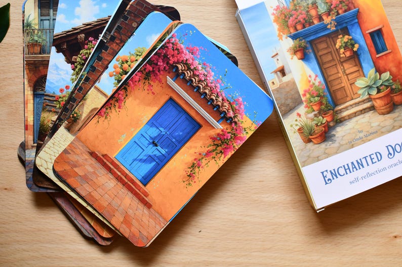 May include: A deck of Enchanted Doors self-reflection oracle cards. The cards feature colourful illustrations of doors and buildings with flowers. The card box is white with blue text and gold accents. The cards are on a wooden surface.
