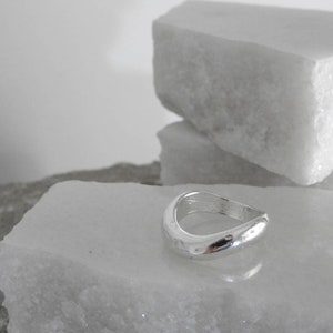 Op de afbeelding: Een zilveren ring met een glad, gebogen ontwerp. De ring ligt op een wit marmeren oppervlak.